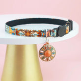 A vibrant orange Bohemian print cat safety collar shown with an attached metal bell and a secure flush-fitting silicone AirTag tracking vault.