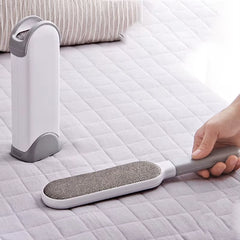 A grey reusable pet hair remover brush being used on a light-colored bed surface, with a matching self-cleaning base shown in the background.