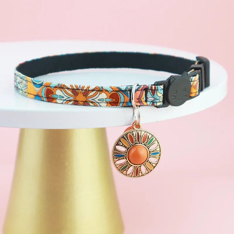 A vibrant orange Bohemian print cat safety collar shown with an attached metal bell and a secure flush-fitting silicone AirTag tracking vault.