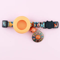 A vibrant orange Bohemian print cat safety collar shown with an attached metal bell and a secure flush-fitting silicone AirTag tracking vault.